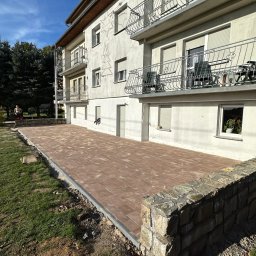 KOLART-BRUK Mykola Chermenskyi - Nowy taras z ceglanej kostki brukowej przy budynku z balkonami, otoczony trawnikiem i kamiennym murkiem. Słoneczny dzień, błękitne niebo.