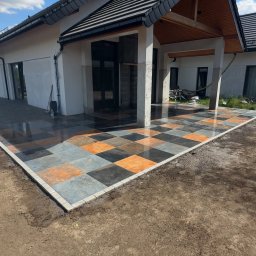 KOLART-BRUK Mykola Chermenskyi - Nowoczesny taras z kostki w szachownicę, obrzeża z jasnej kostki. Dom z ciemnym dachem w tle, błękitne niebo. Taras po deszczu, mokra nawierzchnia.