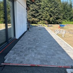 KOLART-BRUK Mykola Chermenskyi - Fragment tarasu z szarej kostki brukowej ułożonej przy domu. Widoczny poziomujący przyrząd, podsypka i elementy krajobrazu. Słoneczny dzień.