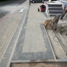 KOLART-BRUK Mykola Chermenskyi - Ułożona kostka brukowa w dwóch odcieniach szarości tworząca wzór na podjeździe. Widoczne schody z kamienia i przygotowane podłoże pod dalsze prace.