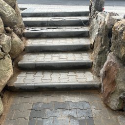 KOLART-BRUK Mykola Chermenskyi - Kamienne schody z kostki brukowej, otoczone dużymi głazami. Widoczne ślady piasku i kabel elektryczny na stopniach. Tło z zielenią drzew.