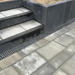 Schody zewnętrzne z szarych płyt kamiennych, obok ułożona kostka brukowa z odpływem liniowym.