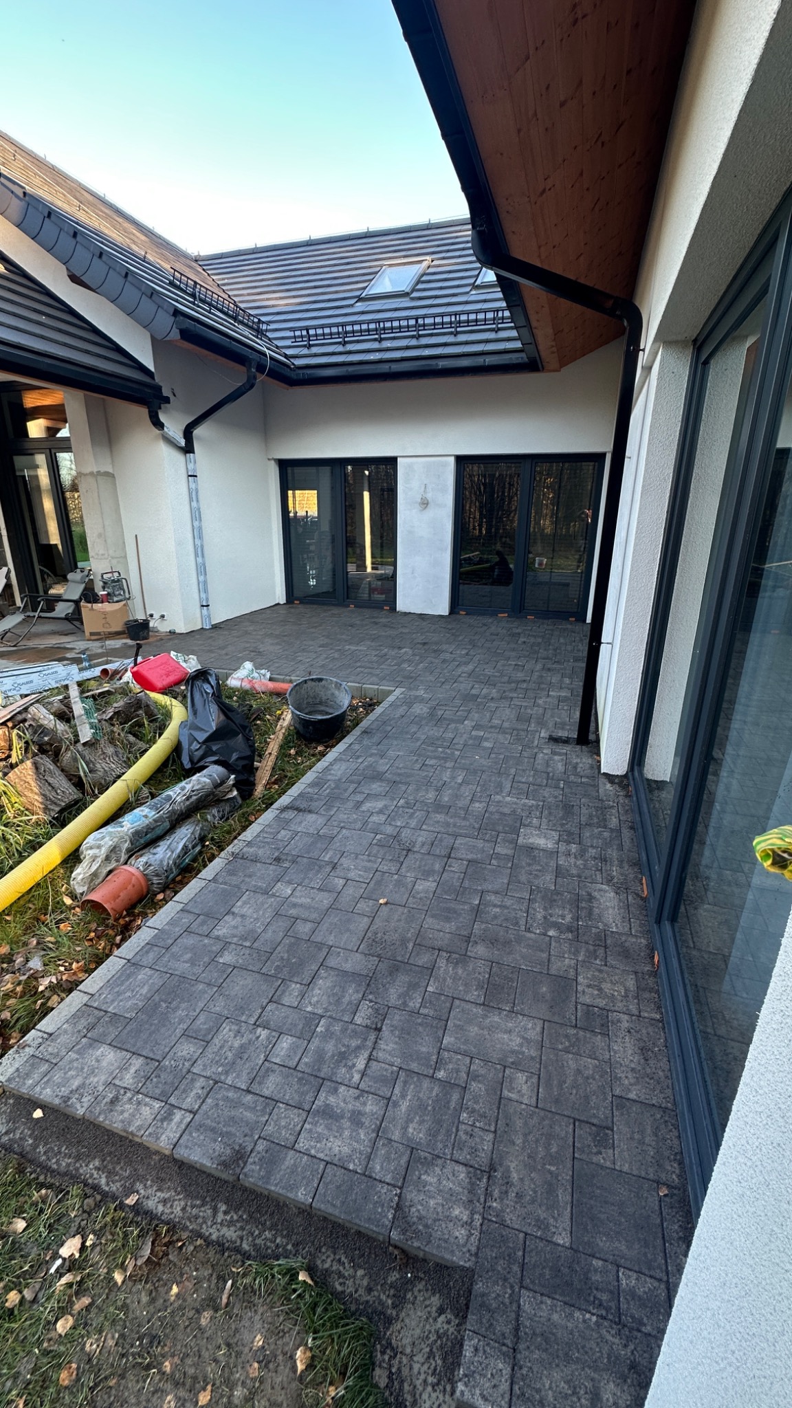 Nowoczesny taras z ciemnoszarej kostki brukowej ułożonej wzorem prostokątnym, z widocznymi elementami wykończeniowymi i narzędziami w tle.