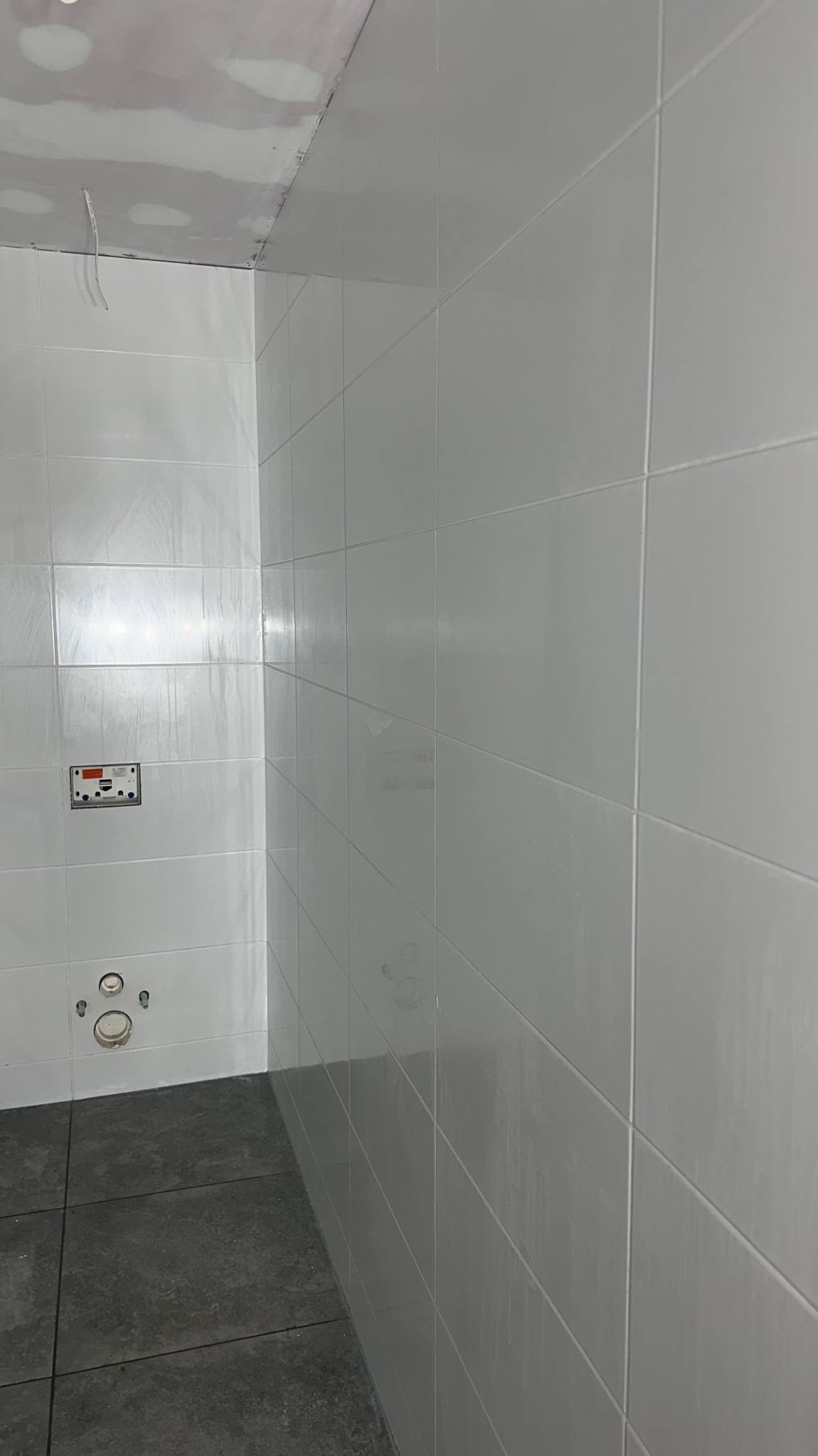 Wykończona łazienka z białymi, błyszczącymi płytkami na ścianach i ciemnoszarą podłogą. Widoczne przyłącza wodno-kanalizacyjne i fragment sufitu w trakcie prac wykończeniowych.