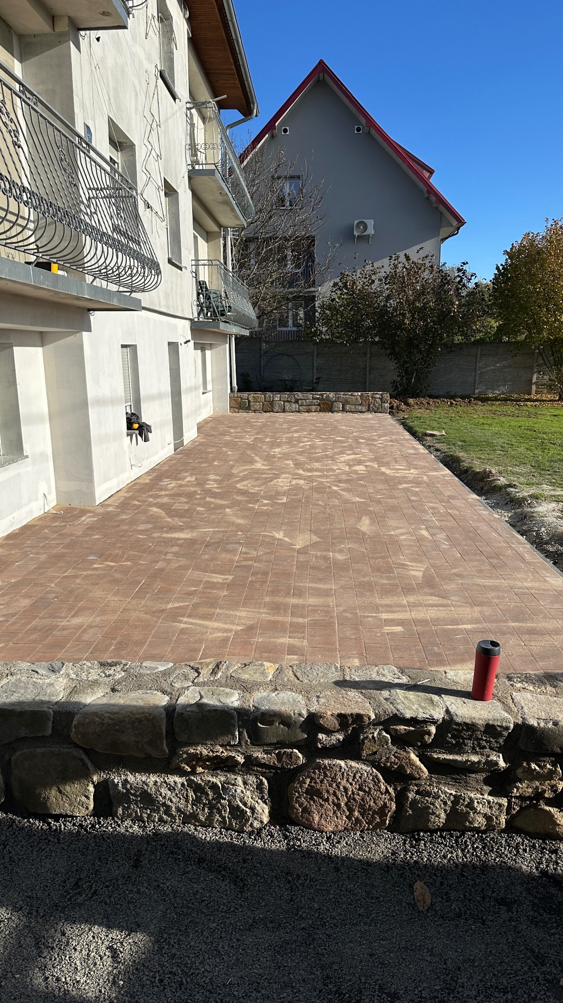 Ułożona, brązowa kostka brukowa na posesji, z kamiennym obrzeżem. Widoczny budynek mieszkalny w tle i fragment ogrodu. Słoneczny dzień.