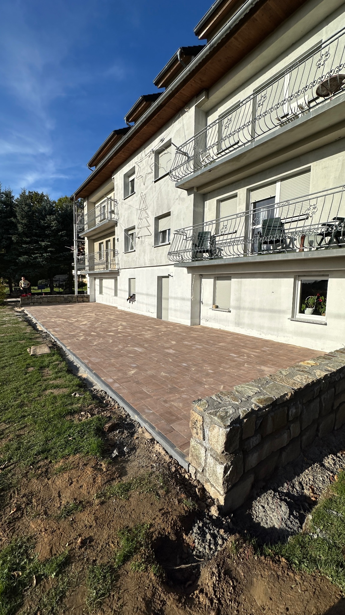 Nowy taras z ceglanej kostki brukowej przy budynku z balkonami, otoczony trawnikiem i kamiennym murkiem. Słoneczny dzień, błękitne niebo.