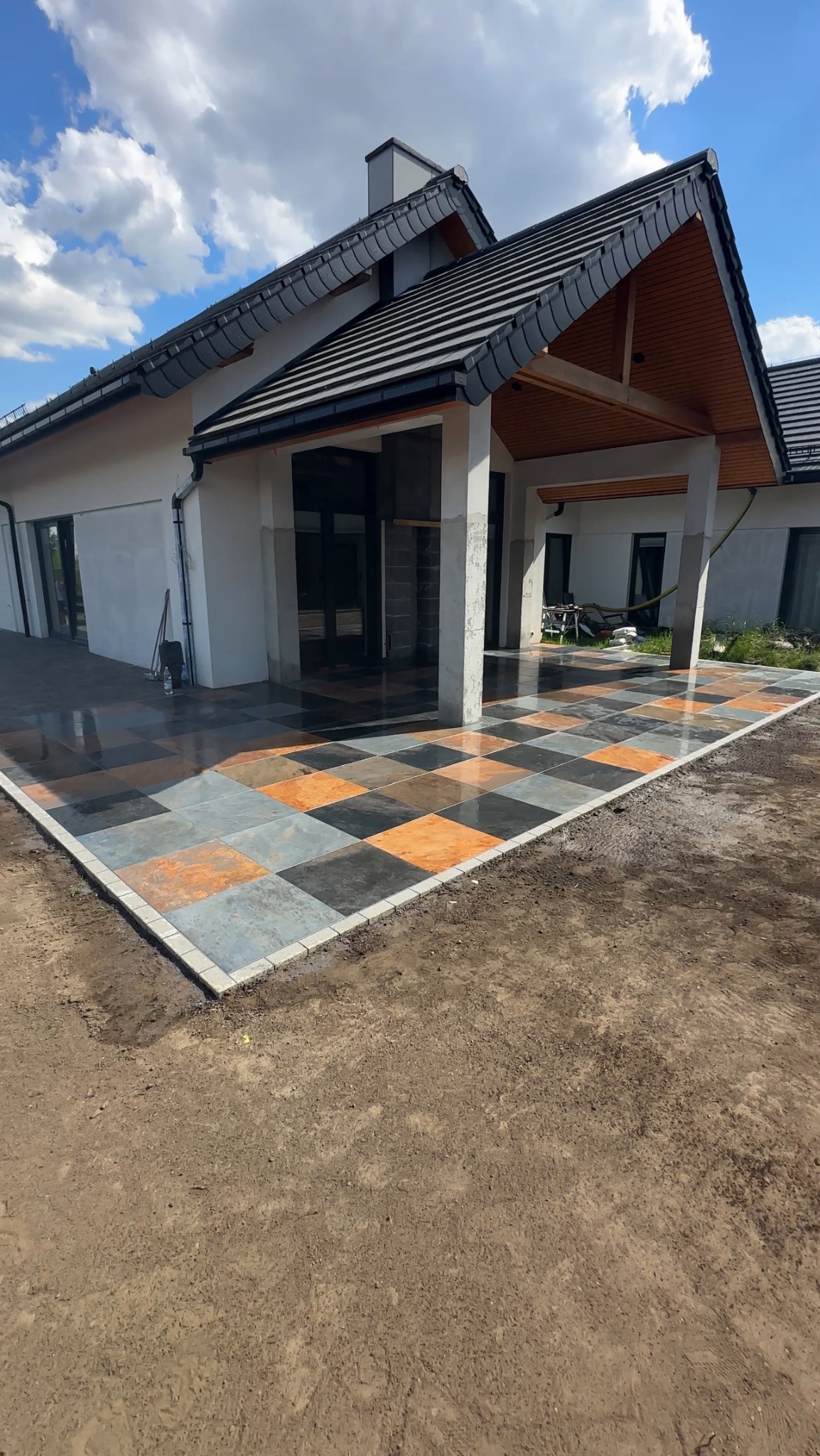 Nowoczesny taras z kostki w szachownicę, obrzeża z jasnej kostki. Dom z ciemnym dachem w tle, błękitne niebo. Taras po deszczu, mokra nawierzchnia.