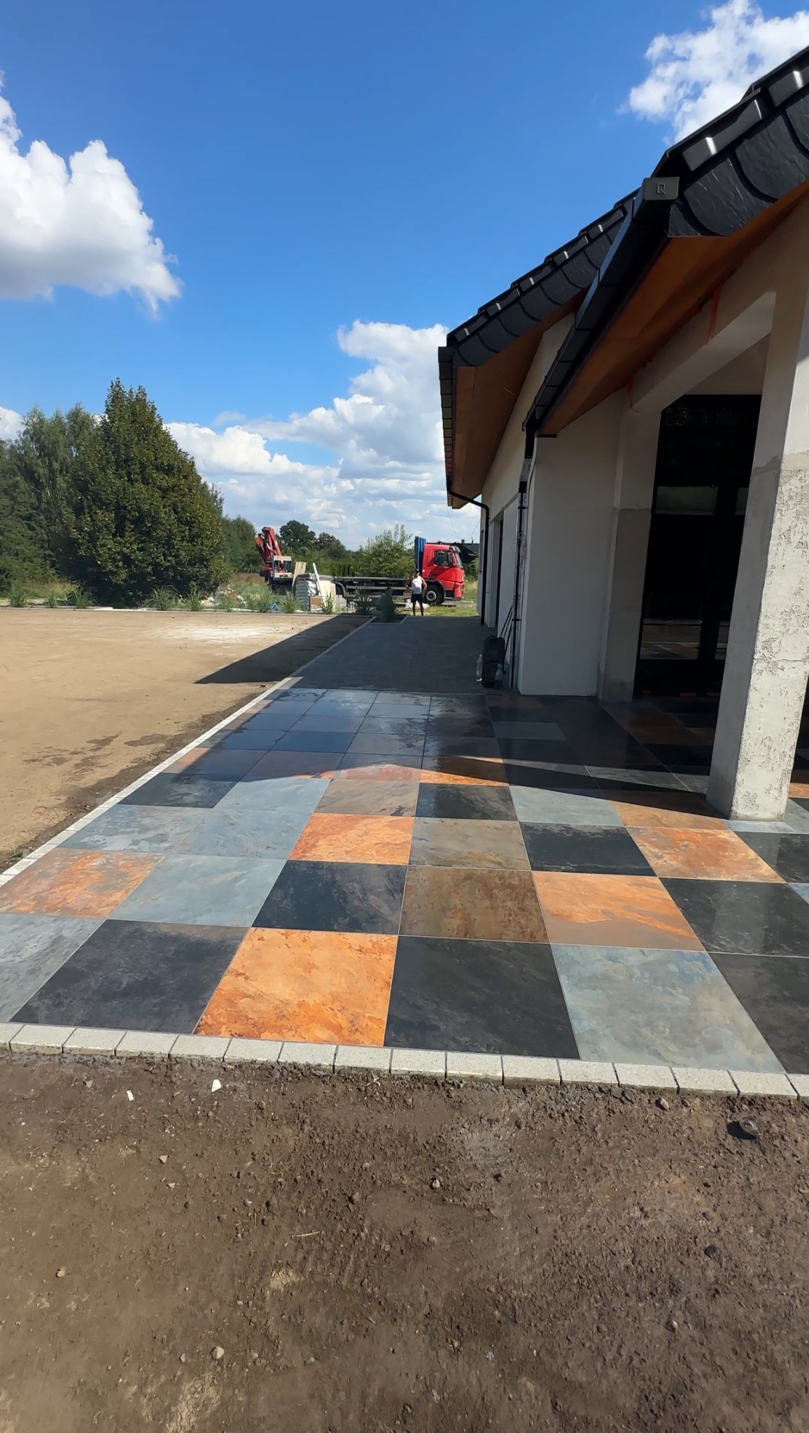 Kolorowa, szachownica z kostki brukowej prowadząca do budynku. W tle widoczny czerwony samochód ciężarowy i maszyny budowlane. Słoneczny dzień.