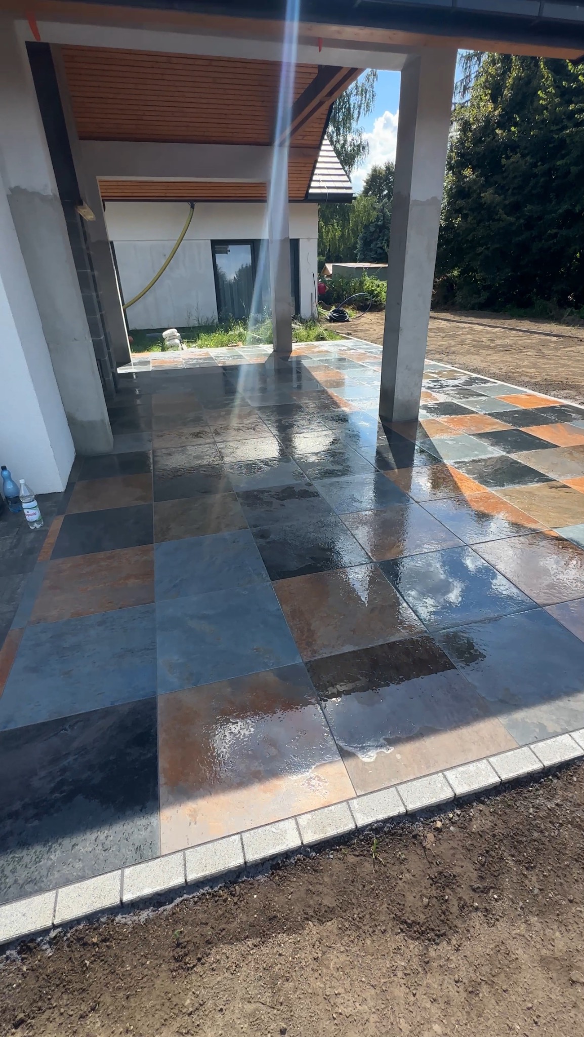 Ułożony taras z kamiennych płyt w odcieniach szarości i brązu, z widocznymi refleksami wody. Krawędź wykończona jasną kostką. W tle dom z drewnianym podbitką.