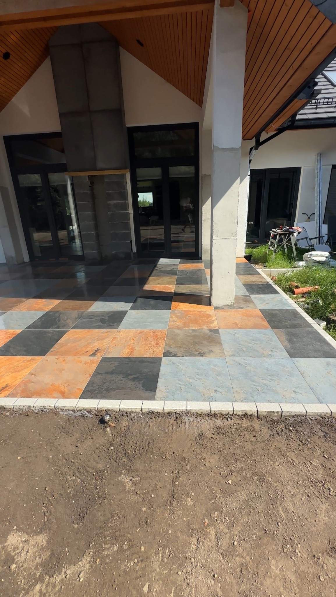 Kolorowa posadzka tarasu z kamiennych płyt ułożonych w szachownicę, obrzeża z jasnej kostki. Widok na dom z dużymi oknami i drewnianym zadaszeniem.