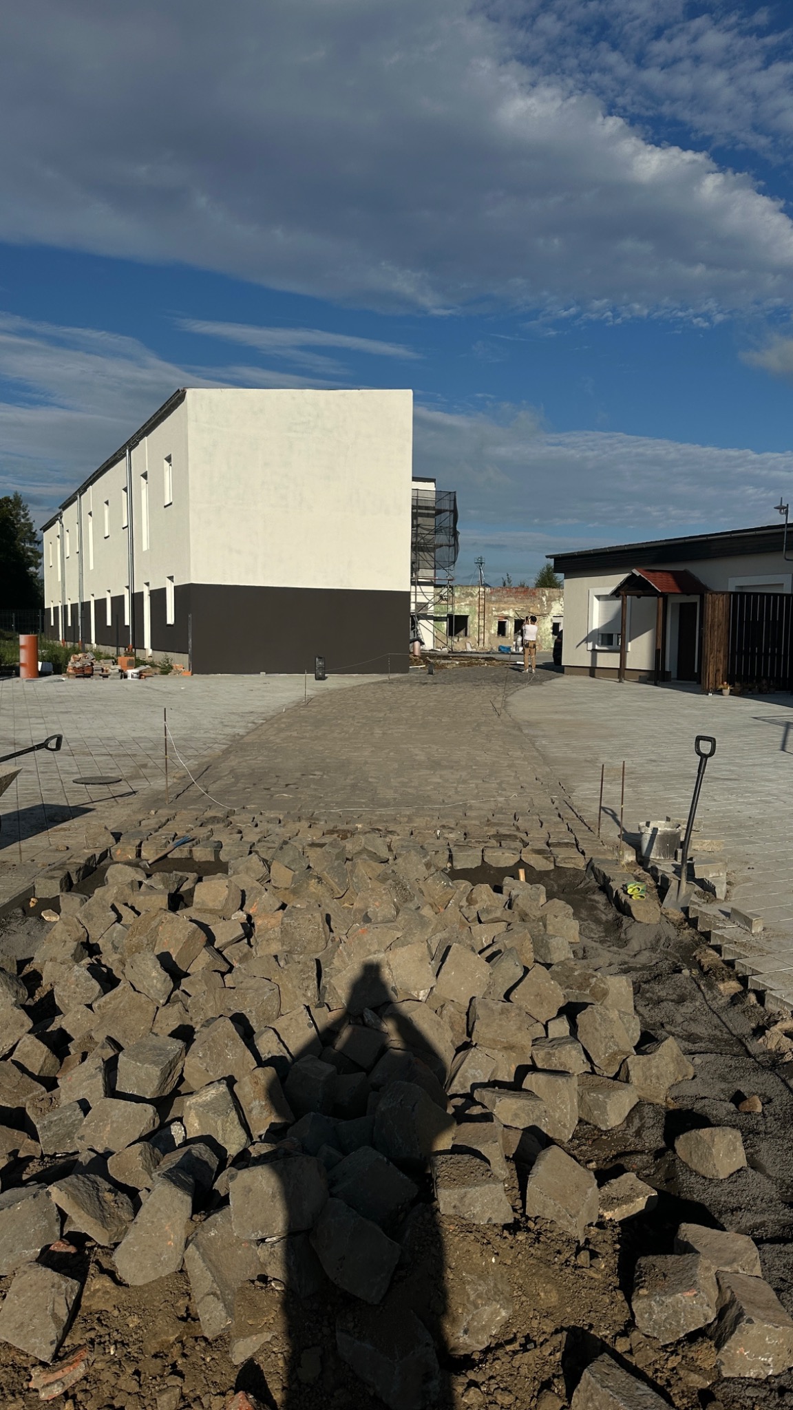 Fragment układania nawierzchni z kostki granitowej na posesji. Częściowo ułożona kostka, w tle budynek i niebo z chmurami. Widoczny cień osoby robiącej zdjęcie.