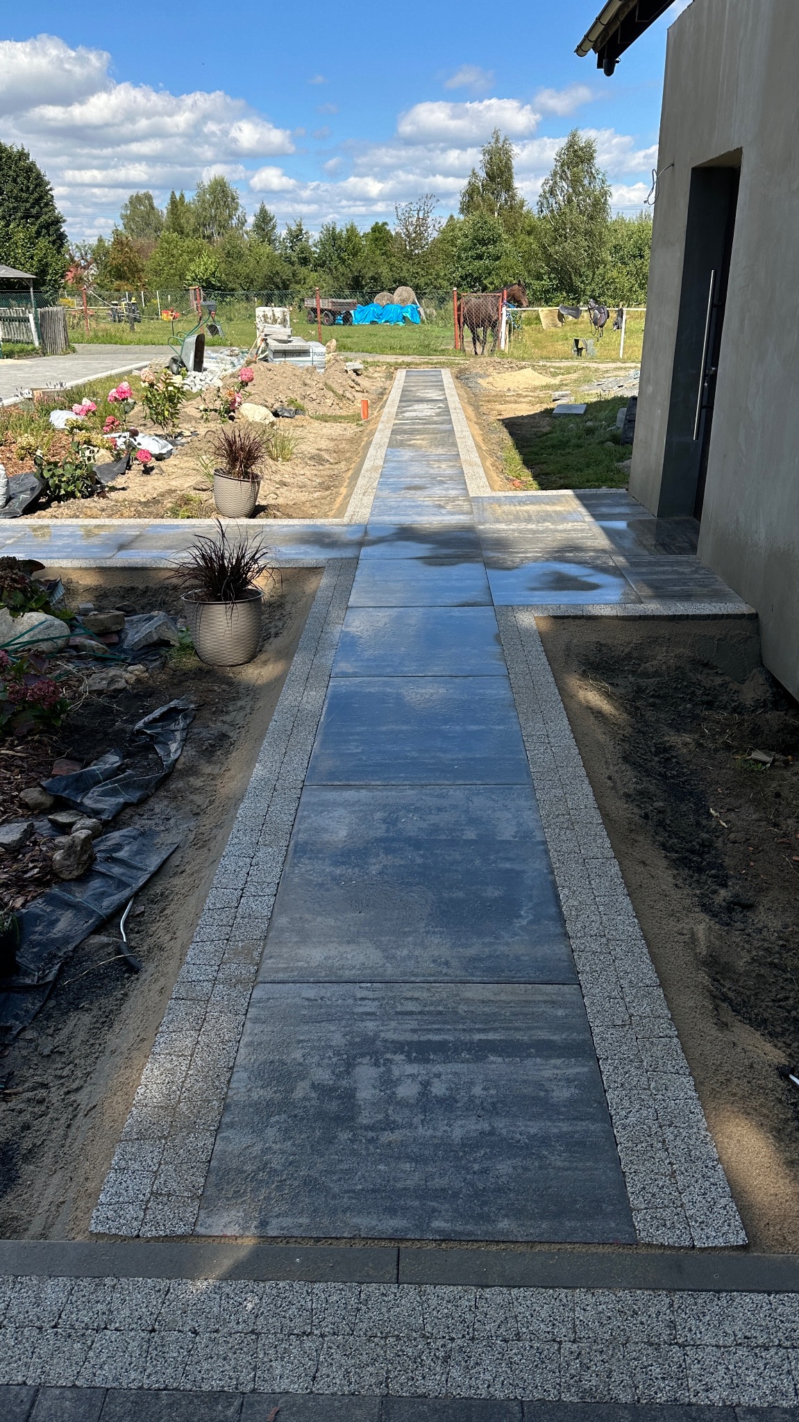 Nowoczesna ścieżka z ciemnej kostki brukowej obramowana jaśniejszą kostką granitową, prowadząca do domu. W tle ogród z roślinami i elementami krajobrazu.