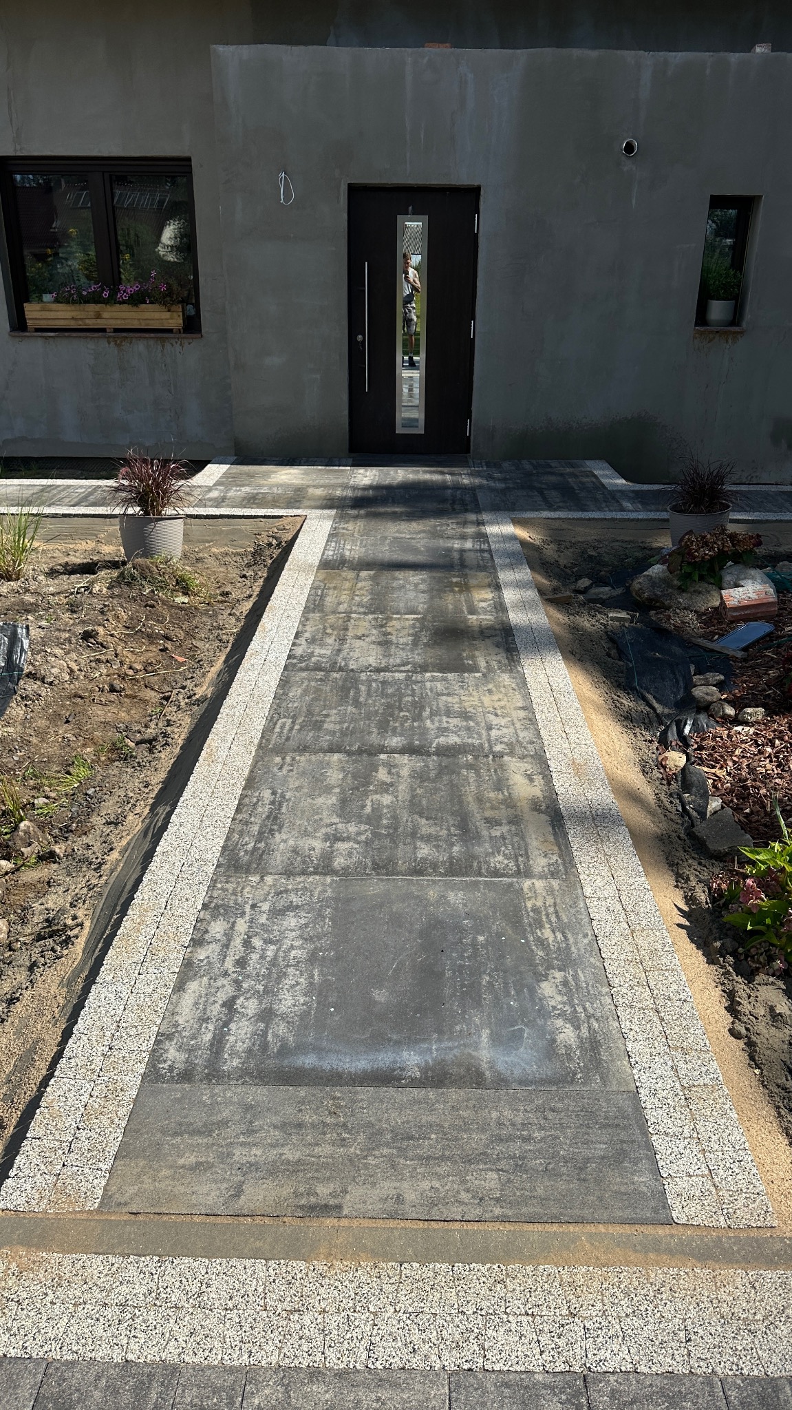 Nowoczesna ścieżka z szarych płyt betonowych obramowana jasną kostką granitową prowadzi do drzwi domu. Widoczne rośliny w donicach i rabatach.