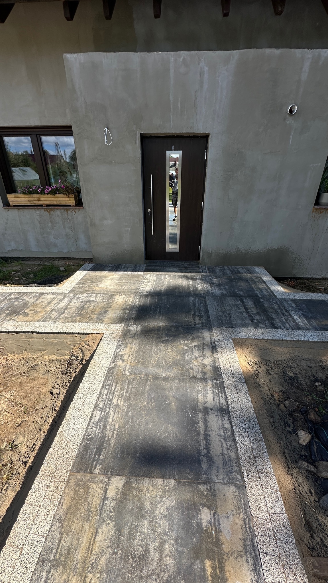 Nowoczesne podejście do domu z szarej kostki brukowej, obrzeża z jasnej kostki granitowej. Widoczne odbicie osoby w drzwiach wejściowych.