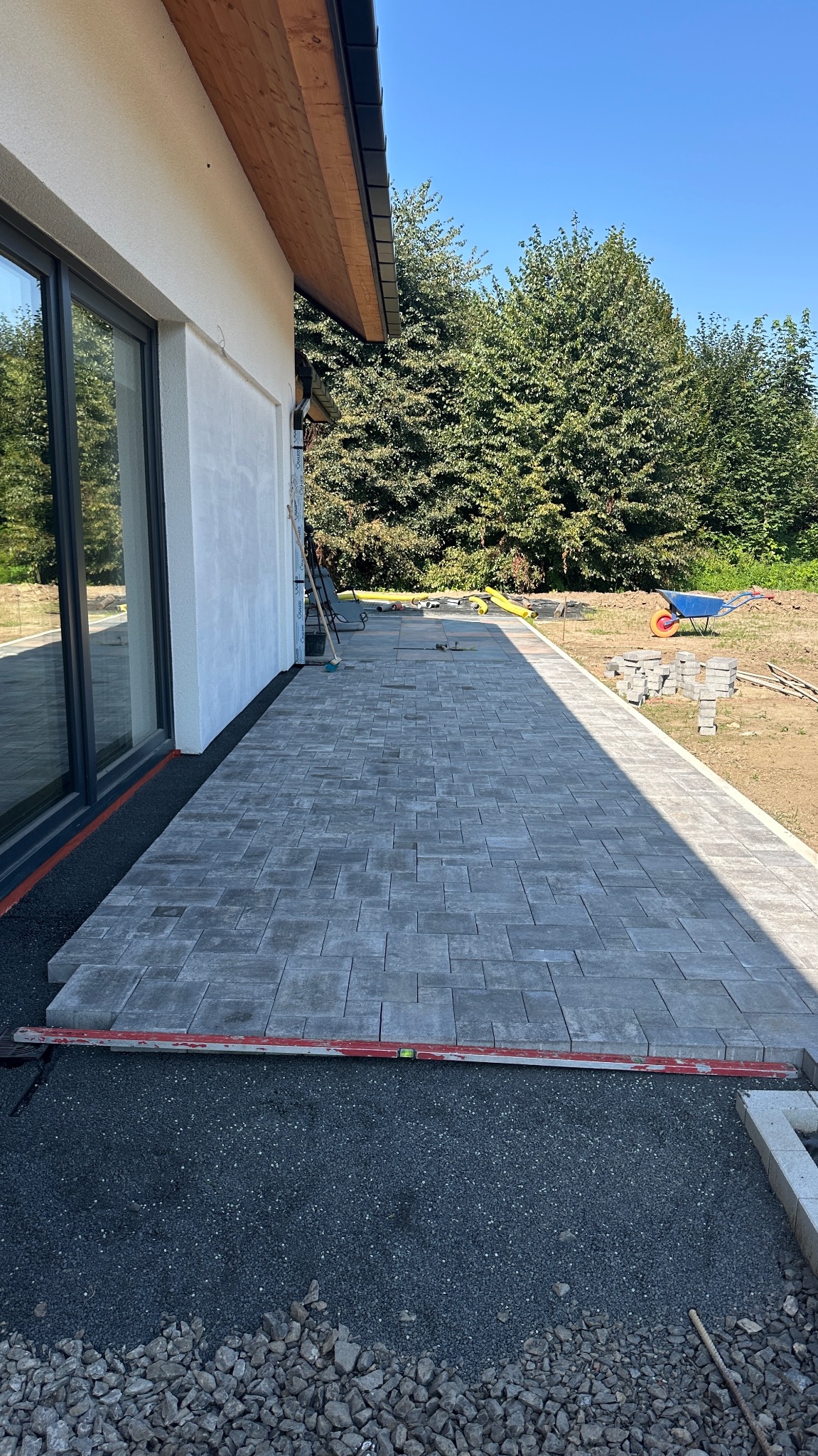 Fragment tarasu z szarej kostki brukowej ułożonej przy domu. Widoczny poziomujący przyrząd, podsypka i elementy krajobrazu. Słoneczny dzień.