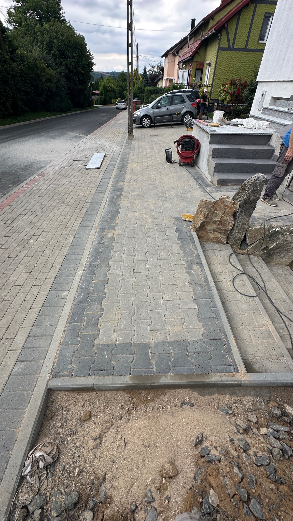 Ułożona kostka brukowa w dwóch odcieniach szarości tworząca wzór na podjeździe. Widoczne schody z kamienia i przygotowane podłoże pod dalsze prace.