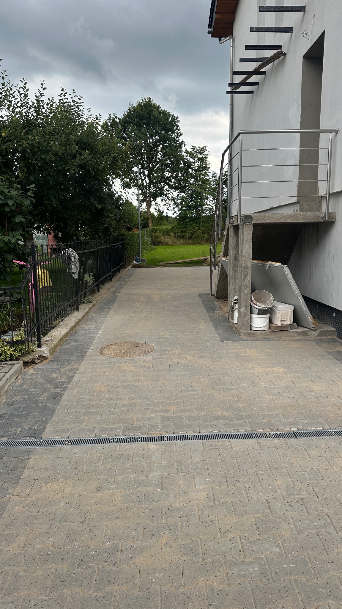 Szara kostka brukowa ułożona na podjeździe, z widocznym odpływem liniowym. Obok domu betonowe schody zewnętrzne z metalową barierką i ogrodzenie z czarnych prętów.