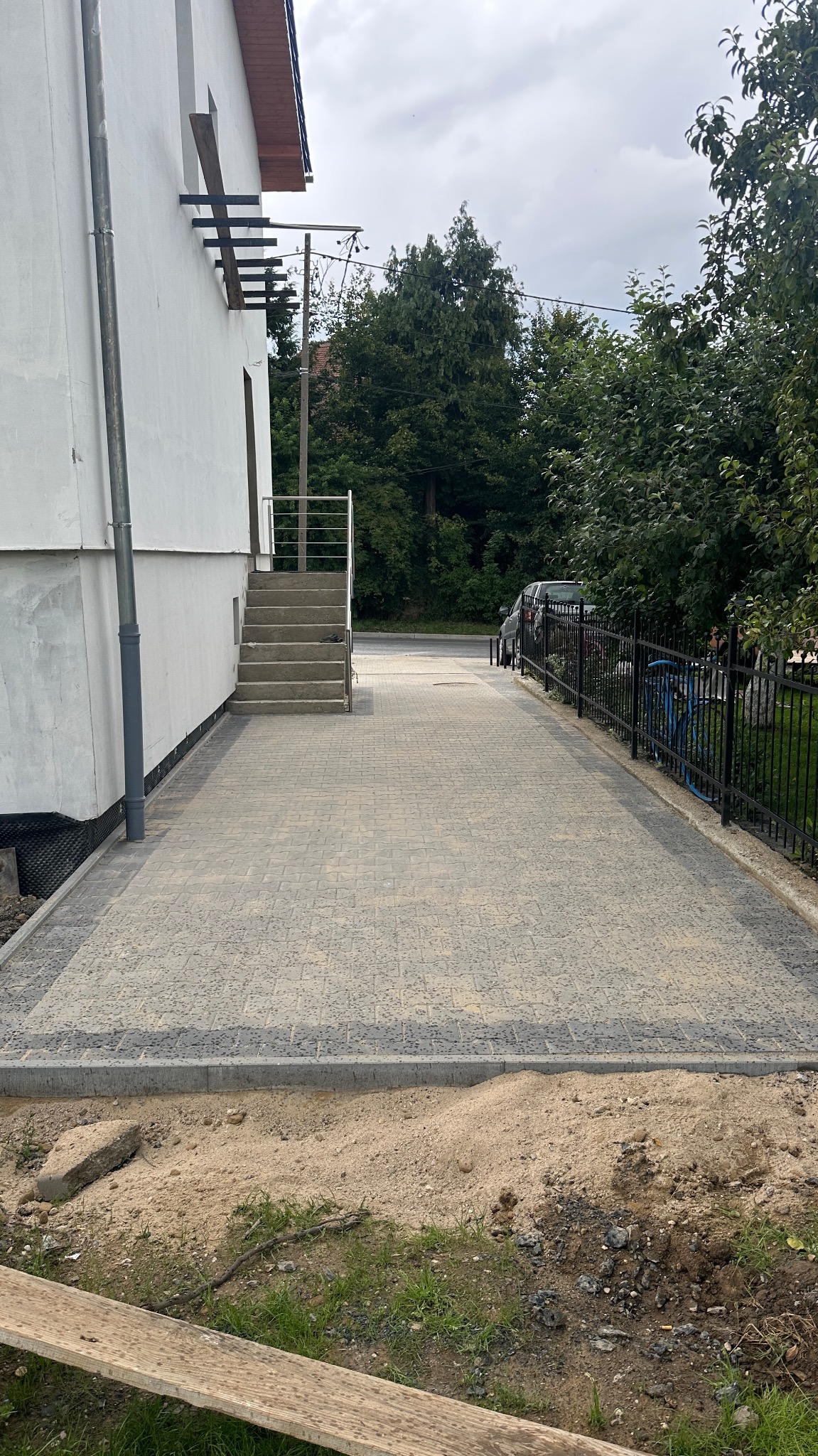 Jasnoszara kostka brukowa ułożona przy budynku z betonowymi schodami i metalową barierką. W tle zieleń drzew i czarne metalowe ogrodzenie.