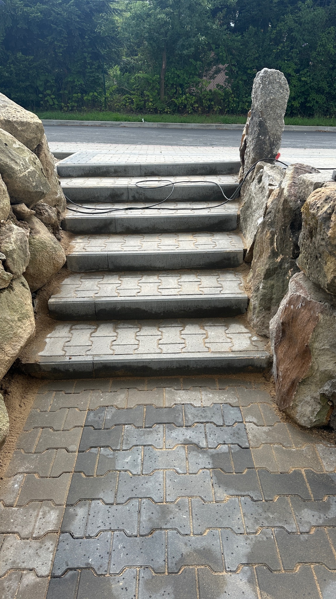 Kamienne schody z kostki brukowej, otoczone dużymi głazami. Widoczne ślady piasku i kabel elektryczny na stopniach. Tło z zielenią drzew.