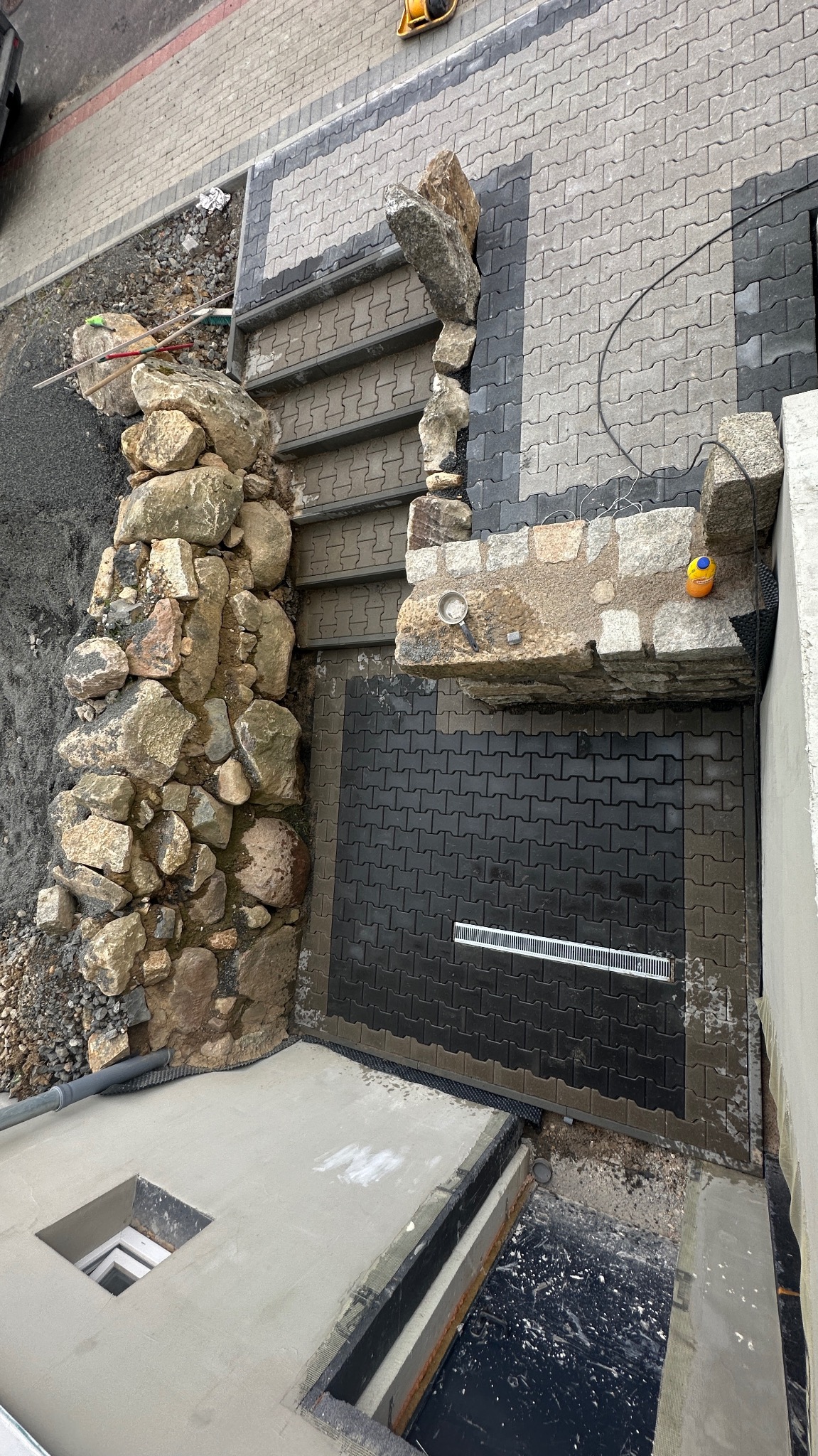 Układanie kostki brukowej i kamiennych schodów z elementami skalnymi. Widok z góry na etap prac z widocznym drenażem liniowym i częściowo ułożoną kostką.