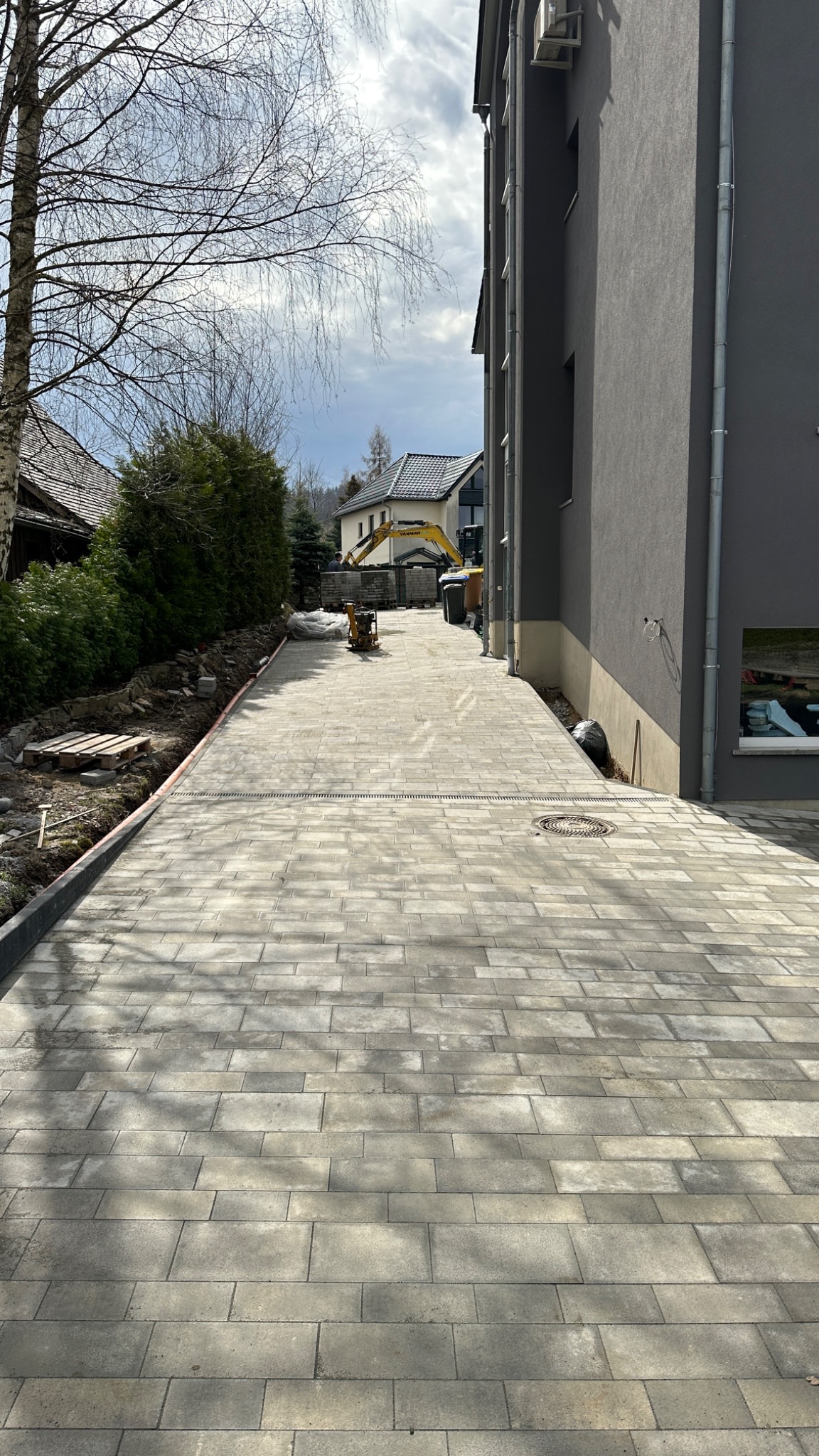 Ułożona szara kostka brukowa prowadząca wzdłuż budynku, z widocznym minikoparką w tle i przygotowanym terenem pod dalsze prace.