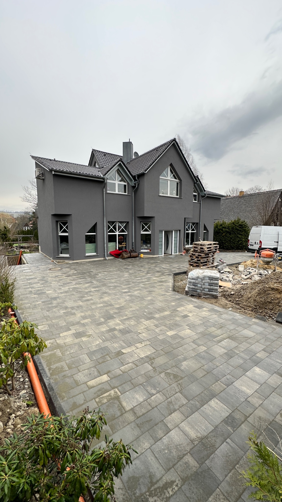 Nowoczesny dom z szarą elewacją i podjazdem z szarej kostki brukowej w trakcie układania, widoczne palety z kostką i narzędzia budowlane.