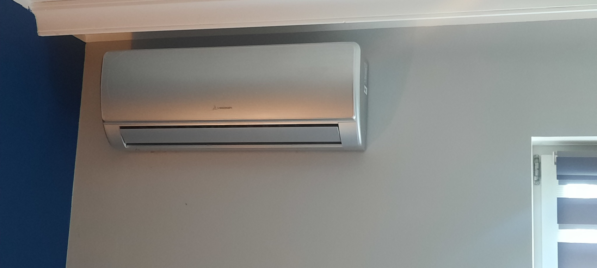 Srebrny klimatyzator Mitsubishi Electric zamontowany na szarej ścianie, widoczny fragment niebieskiej ściany i okno z żaluzjami.