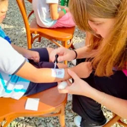 Malowanie tymczasowego tatuażu na ramieniu dziecka za pomocą pędzelka i białego kleju na tle drewnianych krzeseł i żwirowego podłoża.