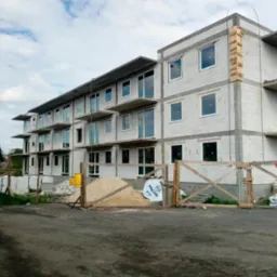 Budynek wielorodzinny w trakcie budowy, z widocznymi oknami, balkonami, oraz materiałami budowlanymi zabezpieczonymi drewnianym ogrodzeniem tymczasowym.