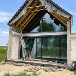 Montaż dużych okien aluminiowych w nowo budowanym domu, widoczne rusztowania i drabina oparta o ścianę, dach w trakcie wykańczania.
