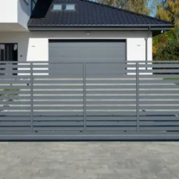 Nowoczesna przesuwna brama wjazdowa w kolorze szarym z poziomymi panelami, zamontowana na białych, betonowych słupkach, prowadząca do domu z garażem i żaluzjami zewnętrznymi na oknach.