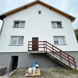 Dwupiętrowy dom z jasną elewacją i ciemnoszarą podmurówką, drewnianymi schodami wejściowymi z brązową balustradą, widoczne puszki z farbą na palecie przy wejściu do piwnicy, pochmurne niebo.