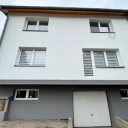 Dwukondygnacyjny dom z nową, białą elewacją i szarym cokołem, widoczne okna z żaluzjami, skrzynka elektryczna z ostrzeżeniem i częściowo odsłonięta boczna ściana budynku.