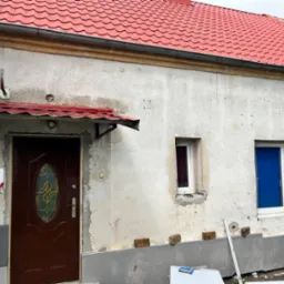 Dom jednorodzinny w trakcie ocieplania elewacji: szara izolacja przy gruncie, czerwona dachówka, okna częściowo zakryte, ślady po starym tynku, przygotowania do dalszych prac.