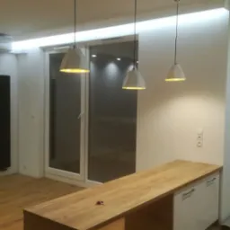 Nowoczesne wnętrze z drewnianym blatem, białymi szafkami, trzema geometrycznymi lampami wiszącymi i oświetleniem LED podsufitowym wzdłuż okna balkonowego.