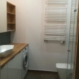 Jasna łazienka z umywalką nablatową, drewnianym blatem, białą szafką, pralką, grzejnikiem drabinkowym i kabiną prysznicową ze szkła.