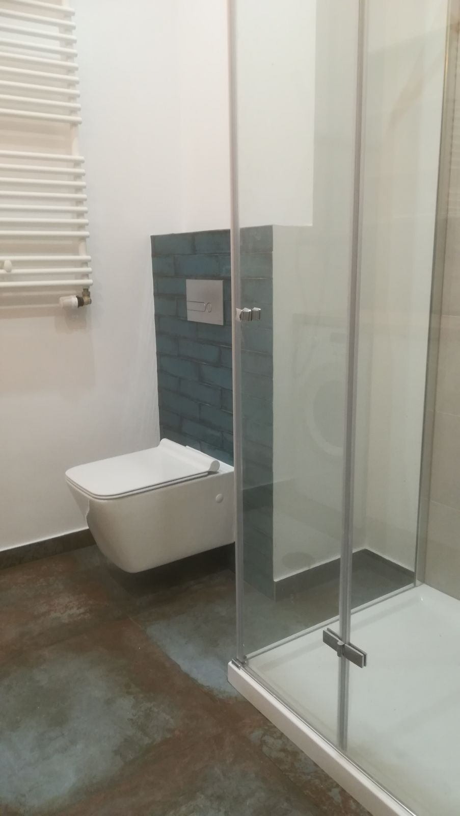 Nowoczesna łazienka z podwieszaną toaletą, niebieską mozaiką za spłuczką i kabiną prysznicową z przezroczystego szkła, widoczny grzejnik drabinkowy.