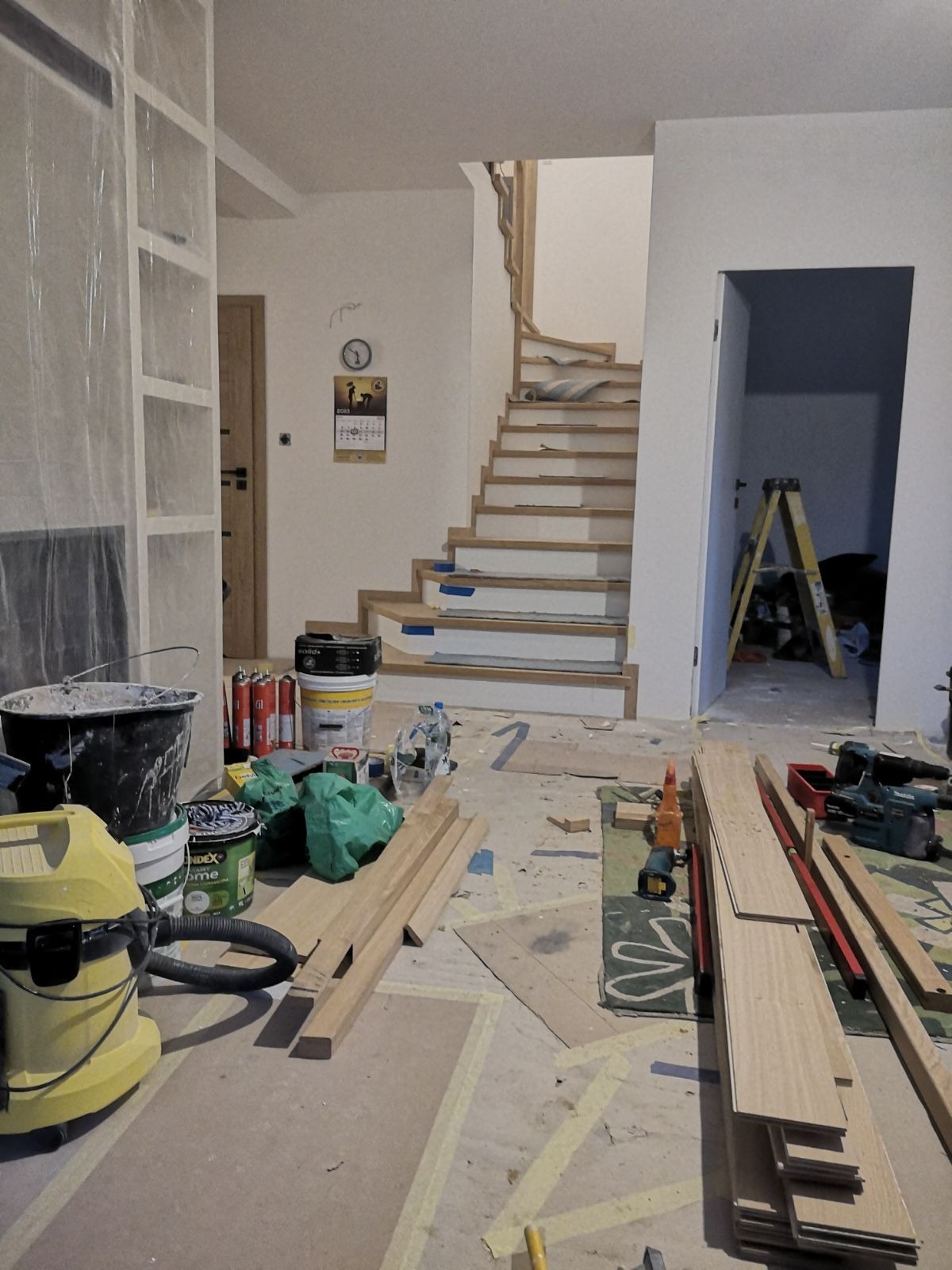 Wnętrze w trakcie remontu: surowe schody drewniane z zabezpieczonymi taśmą krawędziami stopni, narzędzia, materiały budowlane oraz odkurzacz przemysłowy na podłodze zabezpieczonej folią malarską.