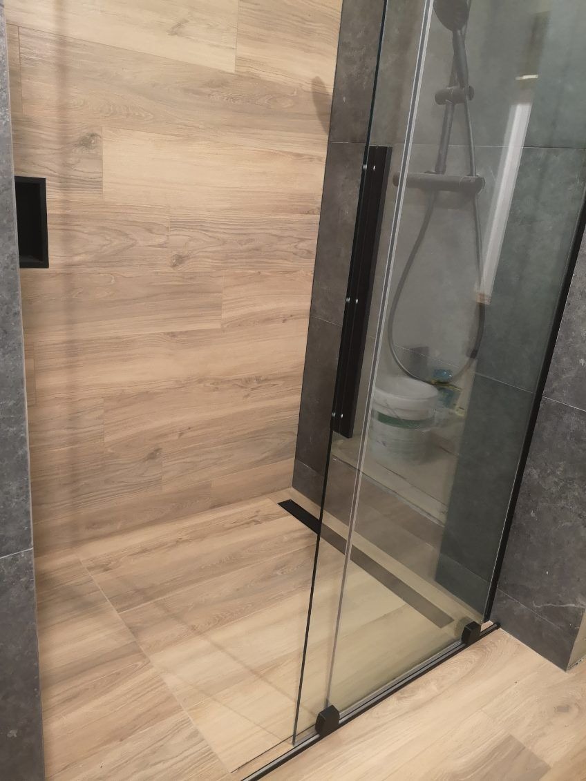 Nowoczesna kabina prysznicowa z przesuwnymi drzwiami, wykończona płytkami imitującymi drewno na ścianie i podłodze, z widocznym odpływem liniowym i fragmentem czarnej wnęki w ścianie.