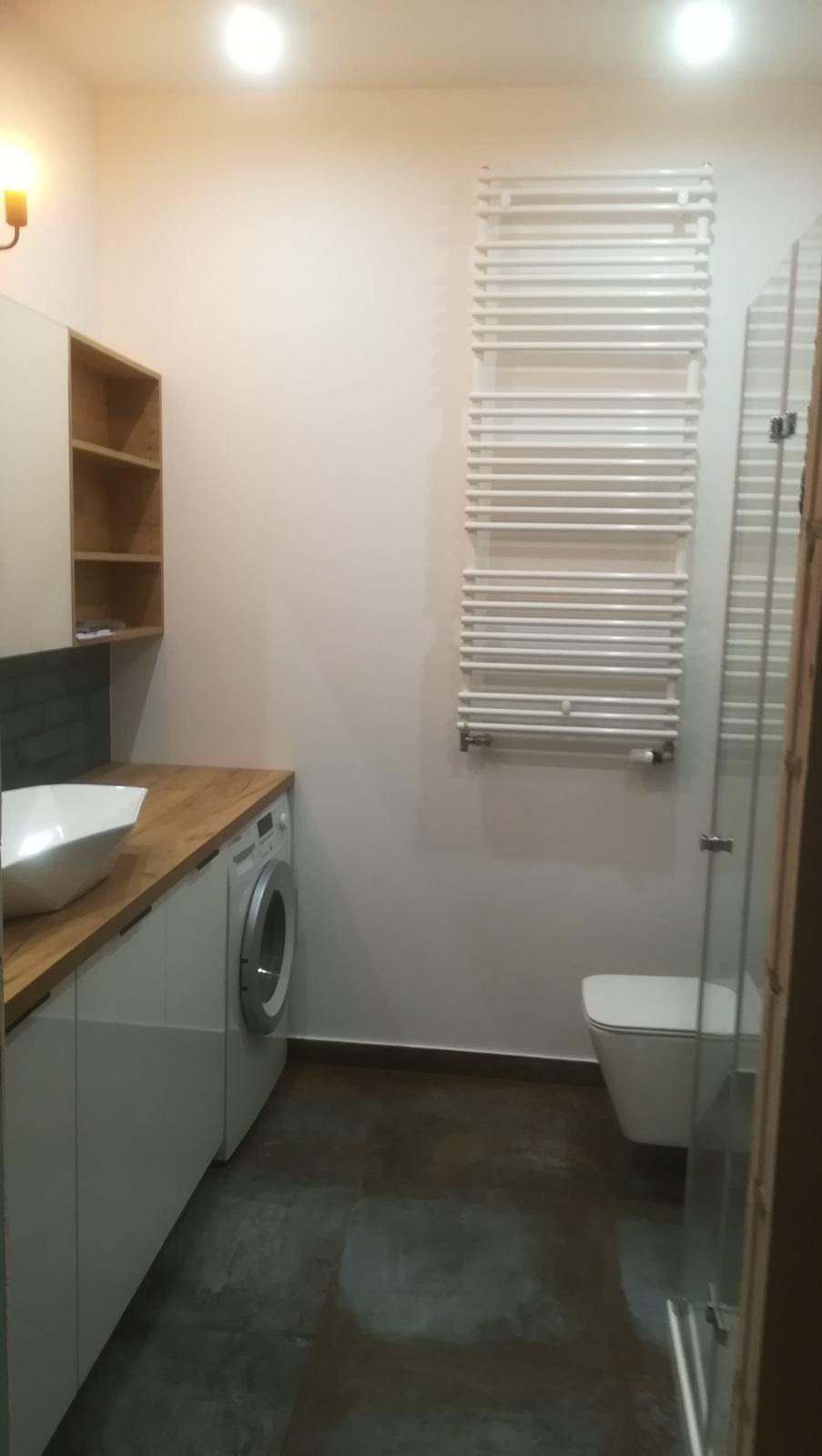 Jasna łazienka z umywalką nablatową, drewnianym blatem, białą szafką, pralką, grzejnikiem drabinkowym i kabiną prysznicową ze szkła.