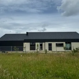 Nowoczesny dom jednorodzinny z ciemną, metalową dachówką na dwuspadowym dachu, widoczny z poziomu trawy, z ogrodzeniem z siatki i młodymi drzewkami w tle.