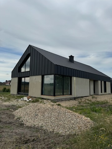 Nowoczesny dom jednorodzinny z czarnym dachem z blachy na rąbek stojący, z dużymi oknami i elewacją w trakcie wykończenia, widoczny stos żwiru przed budynkiem.