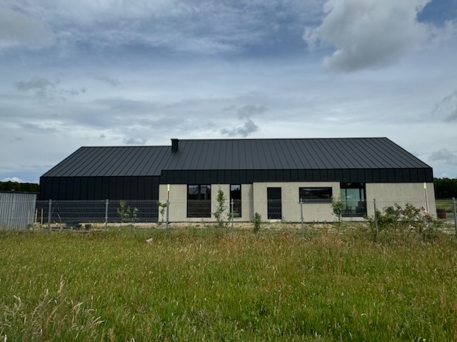 Nowoczesny dom jednorodzinny z ciemną, metalową dachówką na dwuspadowym dachu, widoczny z poziomu trawy, z ogrodzeniem z siatki i młodymi drzewkami w tle.