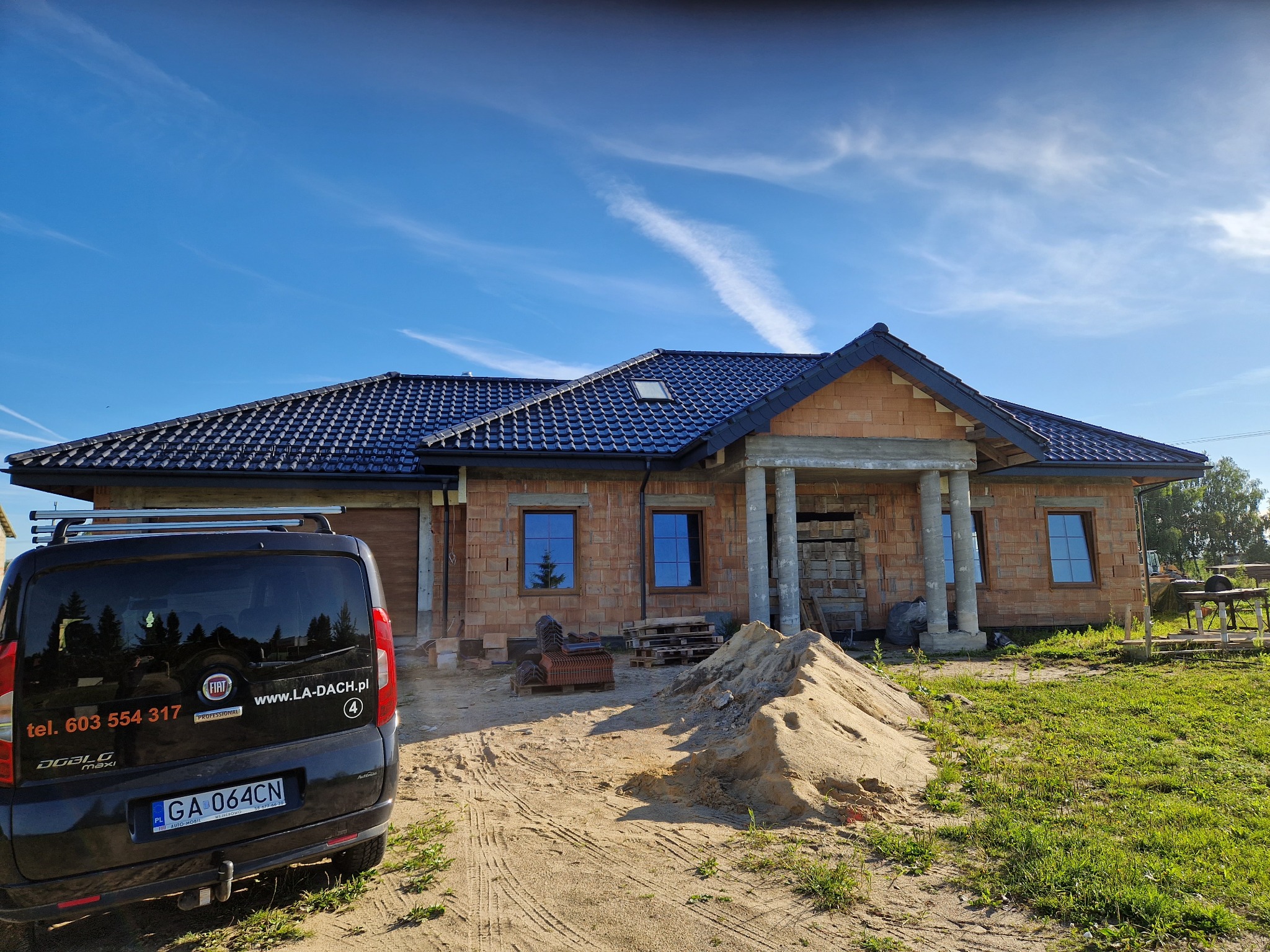 Nowy dom w trakcie budowy z ciemnym, błyszczącym dachem ceramicznym, widoczny samochód firmy dekarskiej z numerem telefonu i adresem strony internetowej, na pierwszym planie piasek i materiały...