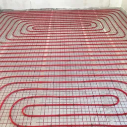 Czerwone rury ogrzewania podłogowego ułożone spiralnie na folii izolacyjnej z siatką, przygotowane do zalania wylewką betonową.