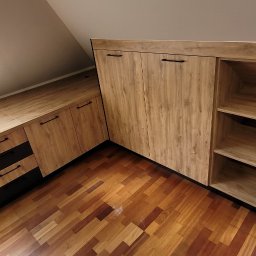 Gggg - Zabudowa pod skosem z drewnianymi frontami i czarnymi detalami, obejmująca szafę z półkami i biurko z szufladami, widoczny parkiet na podłodze.