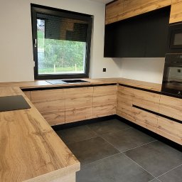 Gggg - Nowoczesna kuchnia z drewnianymi frontami szafek i czarnymi akcentami, widok na okno z roletą i płytę indukcyjną na blacie.