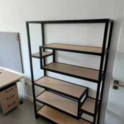 Nowoczesny regał z czarnego metalu z drewnianymi półkami, asymetryczna konstrukcja, widoczny fragment biurka z szufladami w tle.