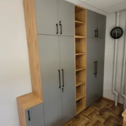 Szara szafa wnękowa z drewnianymi akcentami i otwartymi półkami, zamek w drzwiach, widok na parkiet i rury grzewcze.