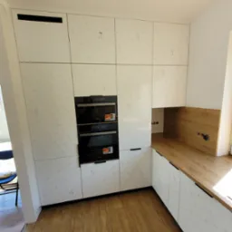 Nowoczesna kuchnia z białymi szafkami z delikatnym wzorem, wbudowanymi piekarnikami i drewnianym blatem, widok z perspektywy podłogi.